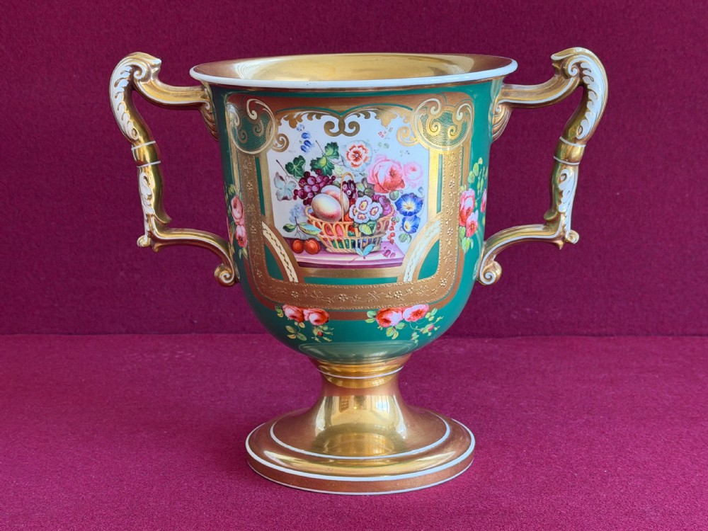 A two-handled green porcelain cup decorated with gold gilding and a hand-painted fruit and floral cartouche on a pedestal foot.
