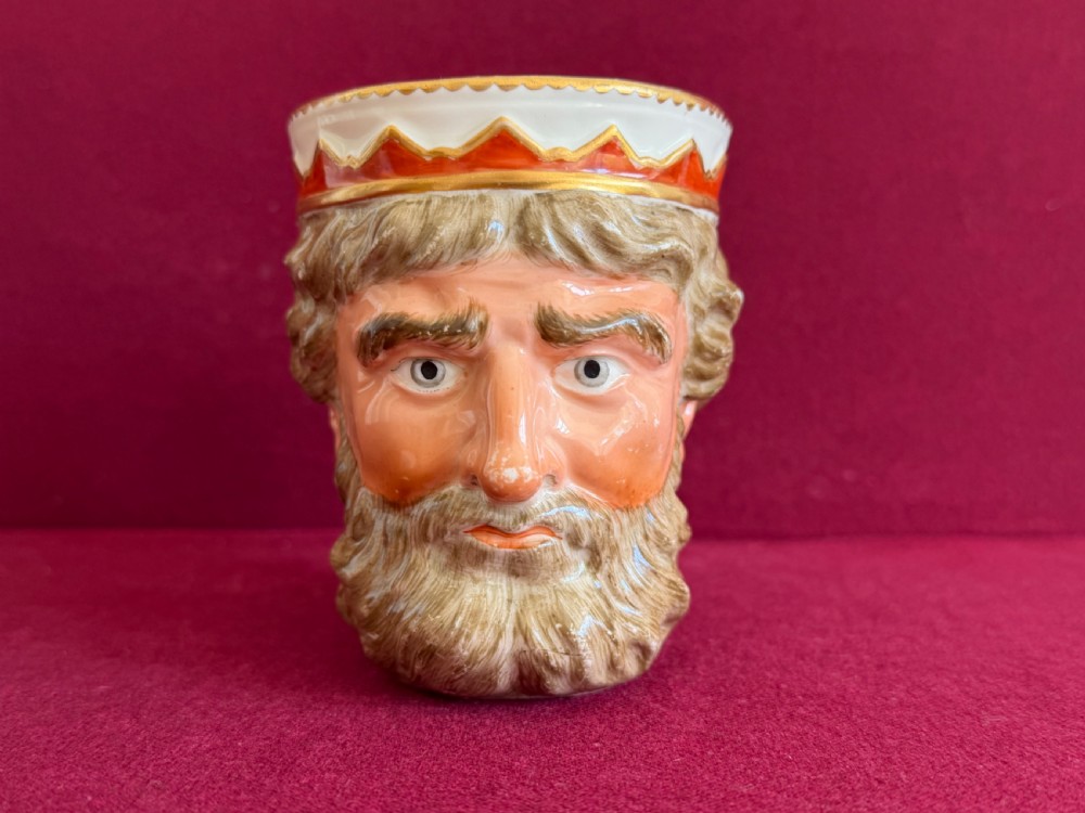 An 18th-century Derby porcelain mug sculpted in the shape of Neptunes head with a painted beard, skin tones, and a red and gold crown rim.