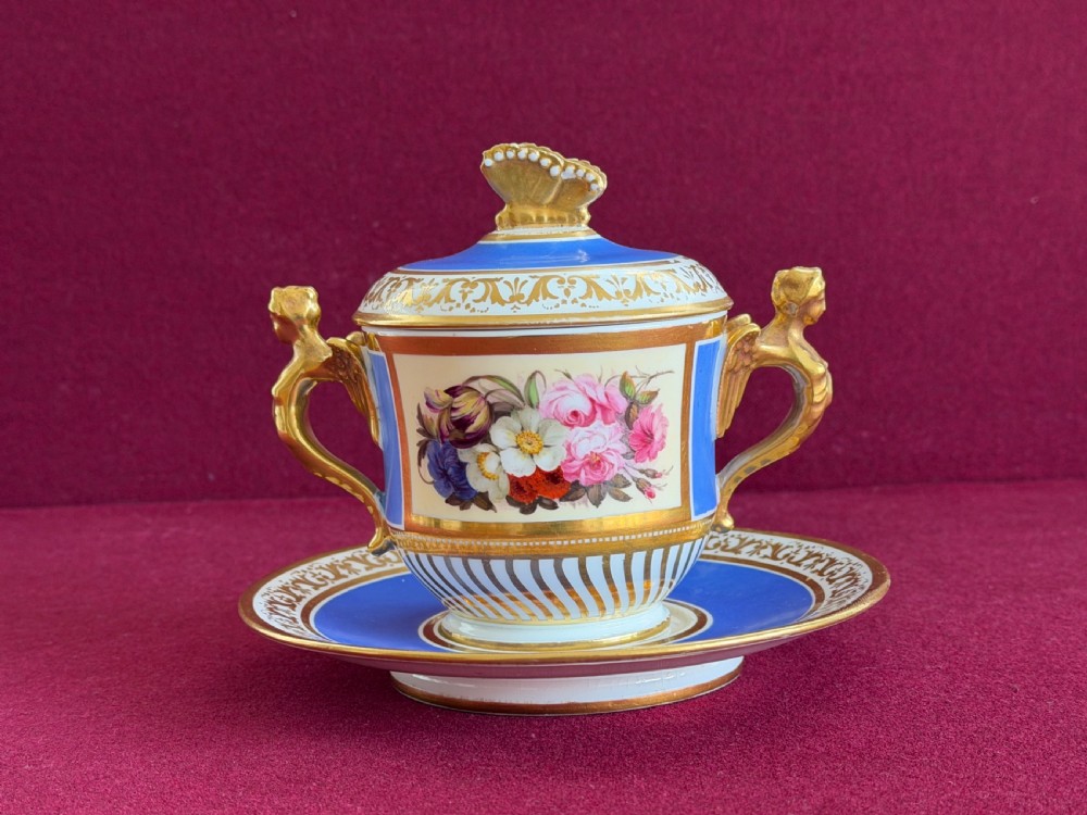 An antique Regency porcelain chocolate cup, cover, and saucer decorated with cobalt blue panels, rich gilding, winged caryatid handles, and a central hand-painted floral bouquet.