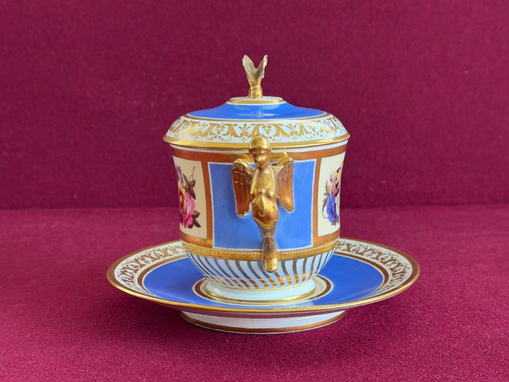 An antique Regency porcelain chocolate cup, cover, and saucer decorated with cobalt blue panels, rich gilding, winged caryatid handles, and a central hand-painted floral bouquet. View II