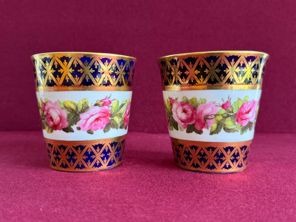 A pair of tapered porcelain beakers featuring a hand-painted floral rose band positioned between cobalt blue borders decorated with gold lattice gilding.