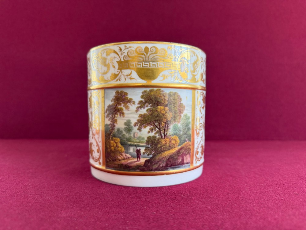 A cylindrical white porcelain porter mug featuring a hand-painted landscape scene framed by ornate gilded scrollwork and classical motifs.