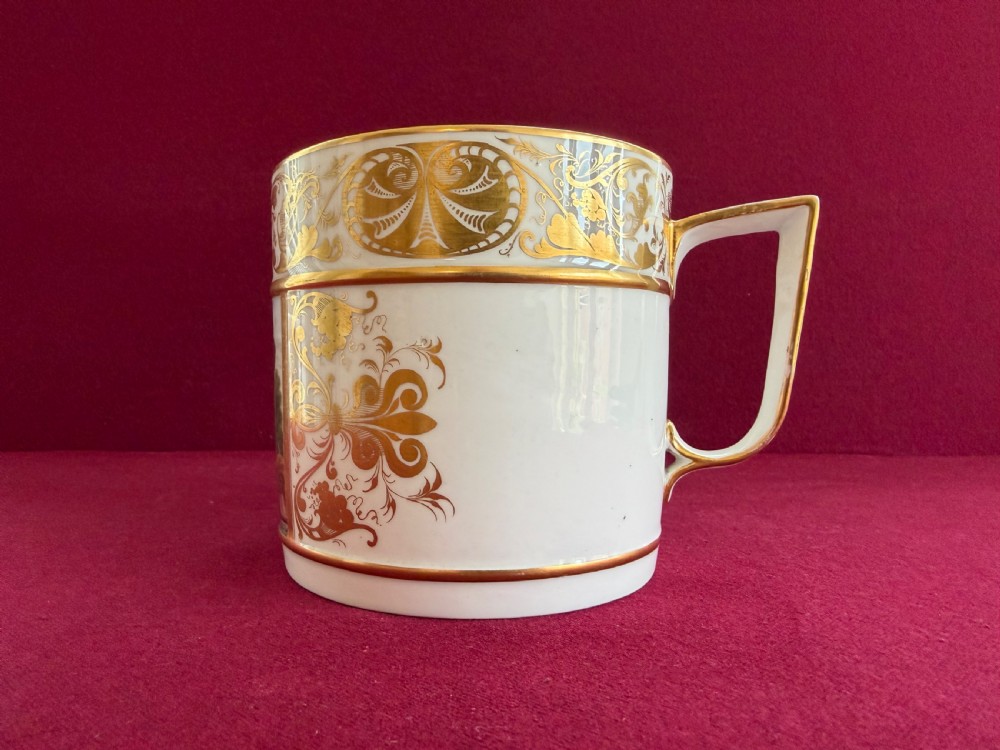A cylindrical white porcelain porter mug featuring a hand-painted landscape scene framed by ornate gilded scrollwork and classical motifs. View II