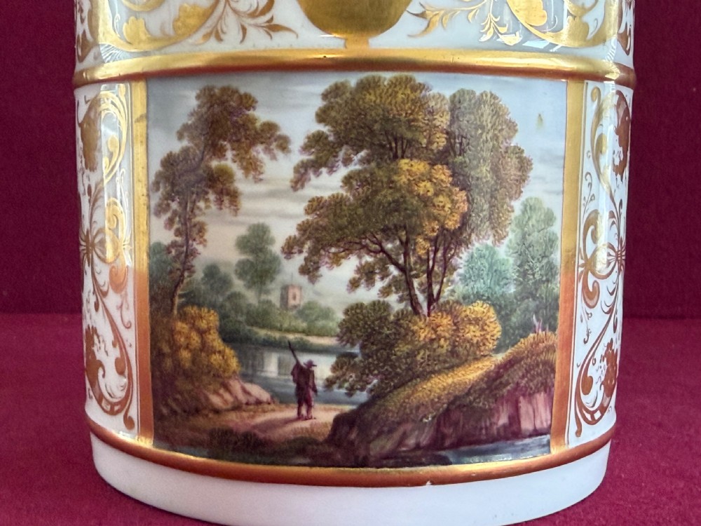 A cylindrical white porcelain porter mug featuring a hand-painted landscape scene framed by ornate gilded scrollwork and classical motifs. View V