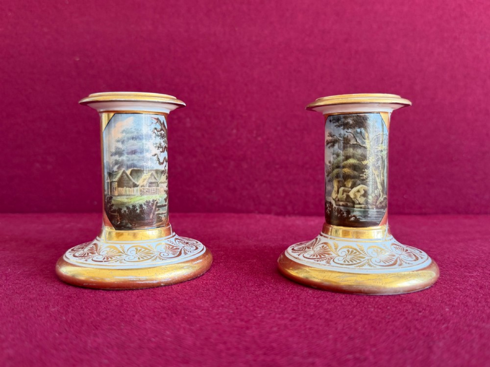 A pair of early nineteenth-century porcelain candlesticks featuring hand-painted landscape scenes on the shafts and detailed gilding on the bases.