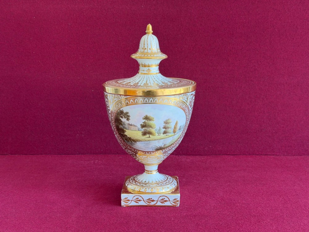 A Neoclassical white porcelain pedestal urn featuring gilded patterns, a domed finial lid, and a central oval reserve painted with a landscape scene.