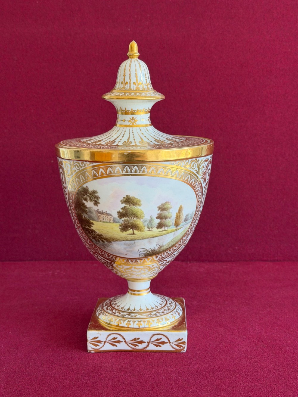 A Neoclassical white porcelain pedestal urn featuring gilded patterns, a domed finial lid, and a central oval reserve painted with a landscape scene. View II