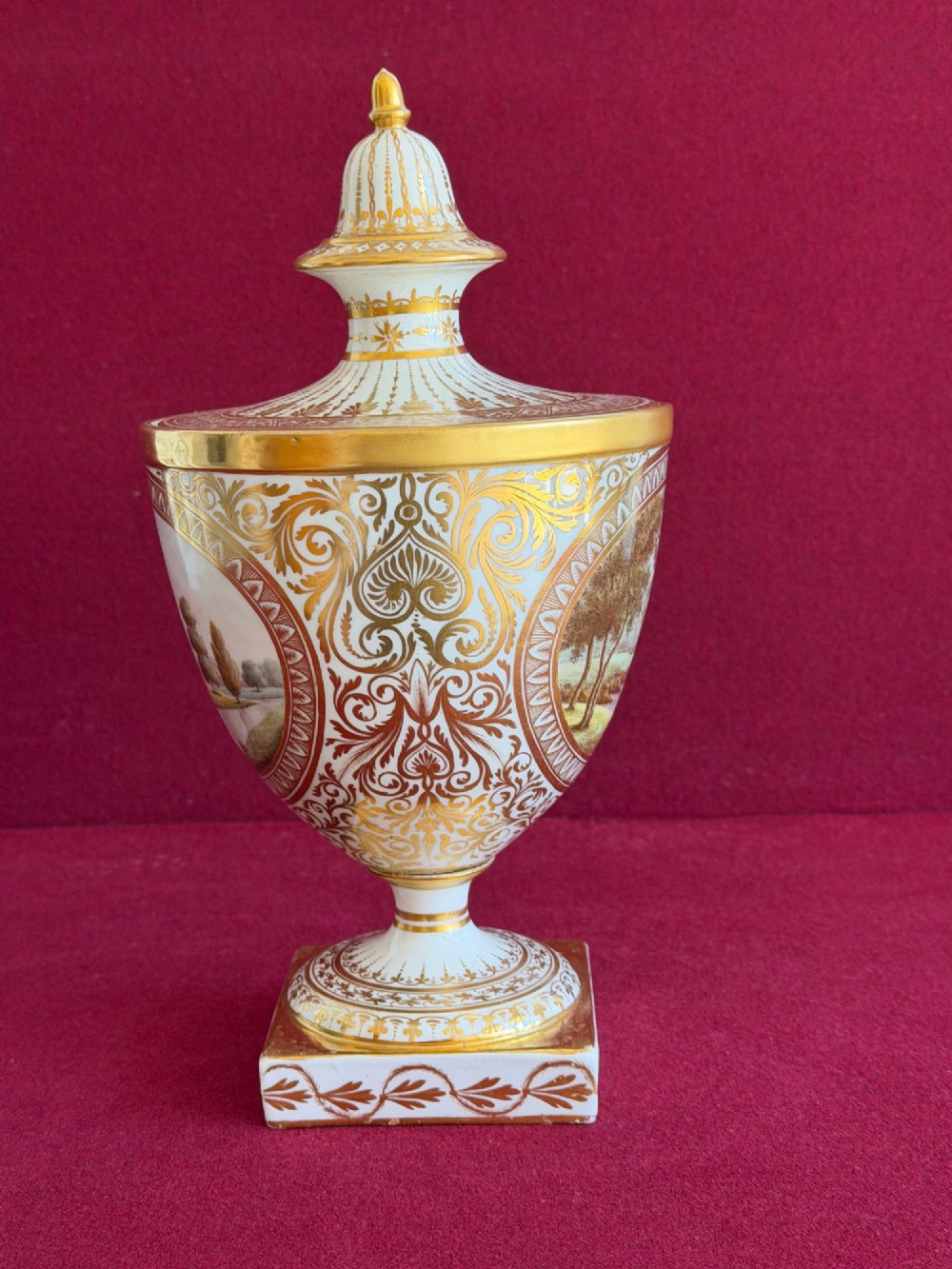 A Neoclassical white porcelain pedestal urn featuring gilded patterns, a domed finial lid, and a central oval reserve painted with a landscape scene. View III