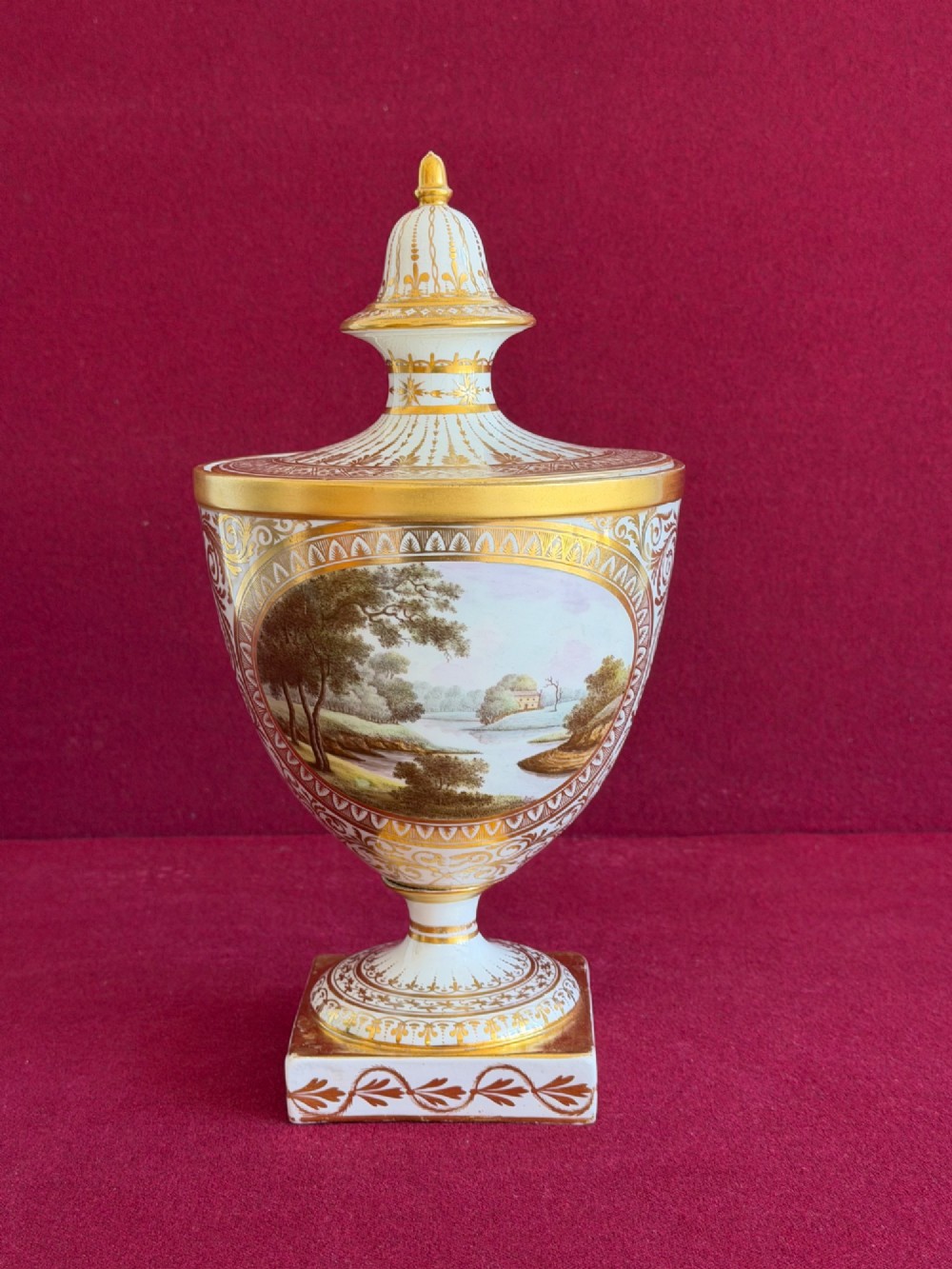 A Neoclassical white porcelain pedestal urn featuring gilded patterns, a domed finial lid, and a central oval reserve painted with a landscape scene. View IV