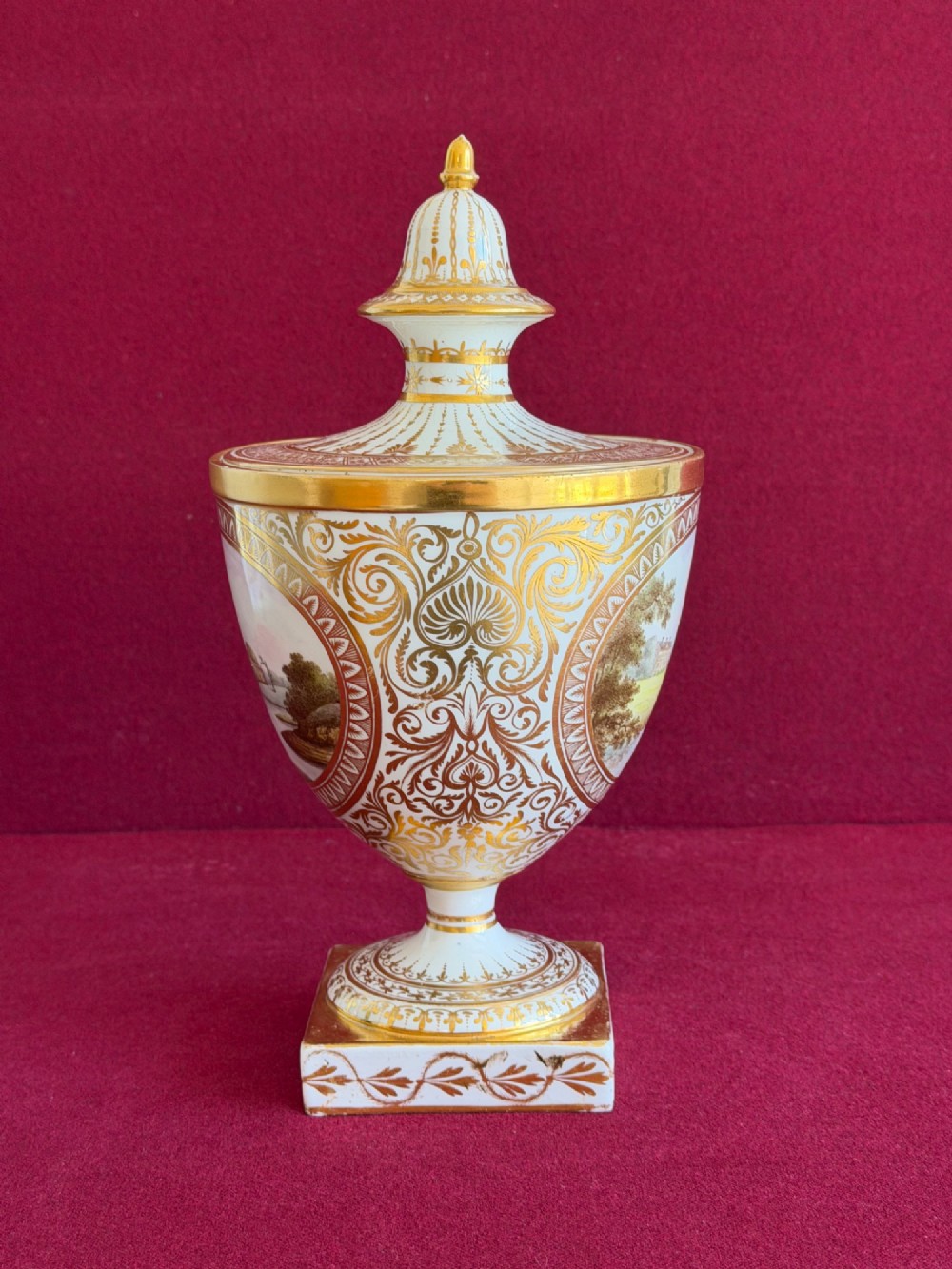 A Neoclassical white porcelain pedestal urn featuring gilded patterns, a domed finial lid, and a central oval reserve painted with a landscape scene. View V