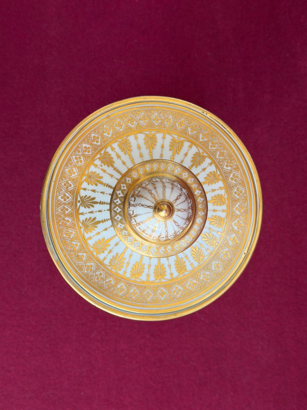 A Neoclassical white porcelain pedestal urn featuring gilded patterns, a domed finial lid, and a central oval reserve painted with a landscape scene. View VI