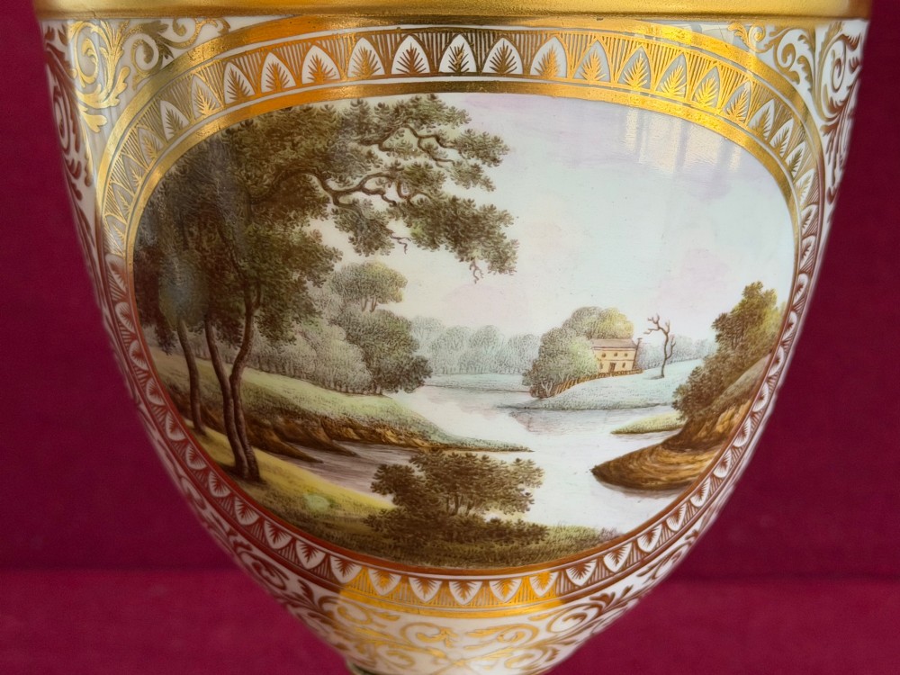 A Neoclassical white porcelain pedestal urn featuring gilded patterns, a domed finial lid, and a central oval reserve painted with a landscape scene. View VII