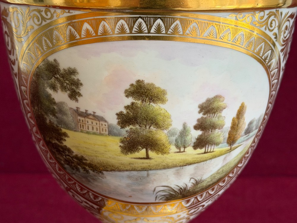 A Neoclassical white porcelain pedestal urn featuring gilded patterns, a domed finial lid, and a central oval reserve painted with a landscape scene. View IX