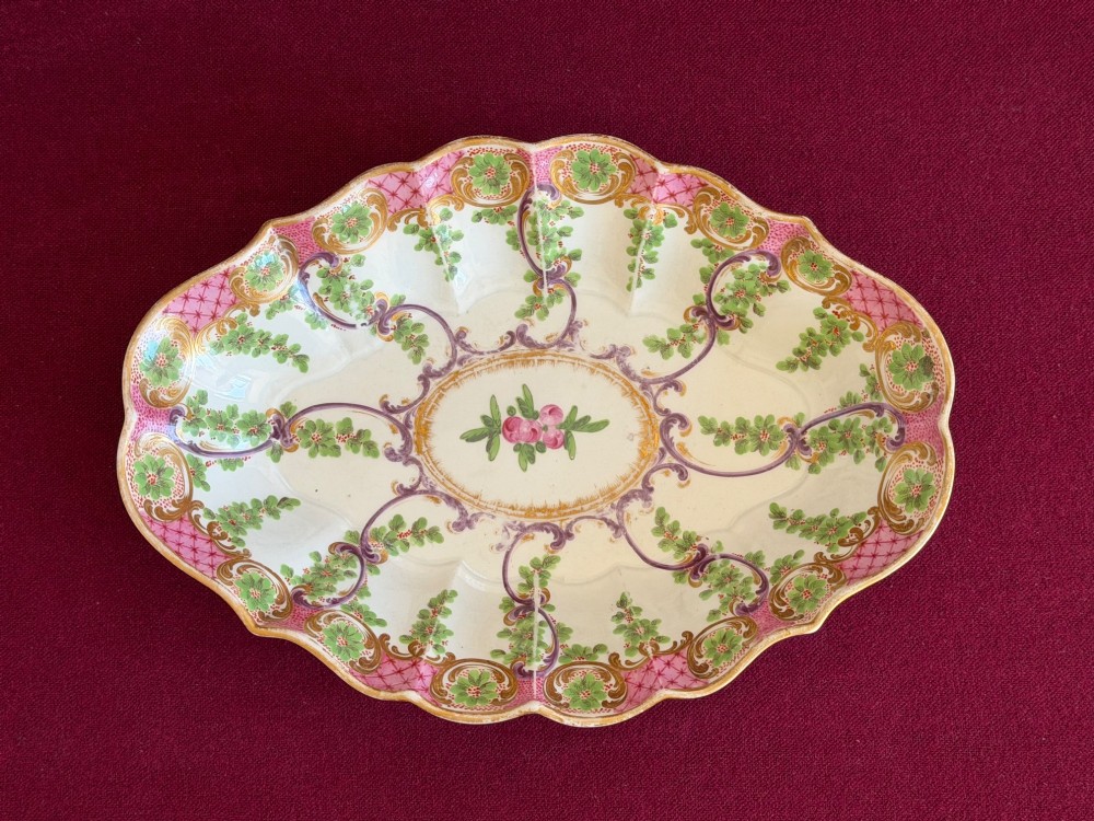 An eighteenth-century oval porcelain dish featuring scalloped edges, pink lattice borders, green foliage garlands, and a central floral motif.