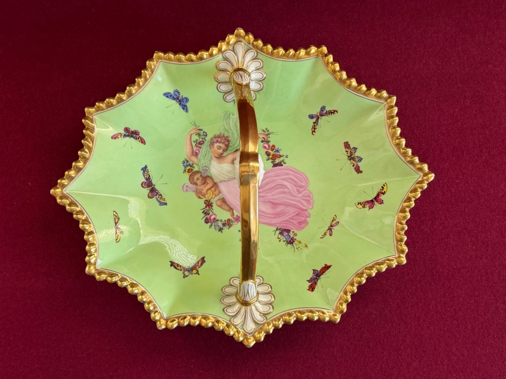 An apple green lobed porcelain basket with a gold overhead handle, gadrooned gilded border, and hand-painted central vignette of a classical figure and butterflies.