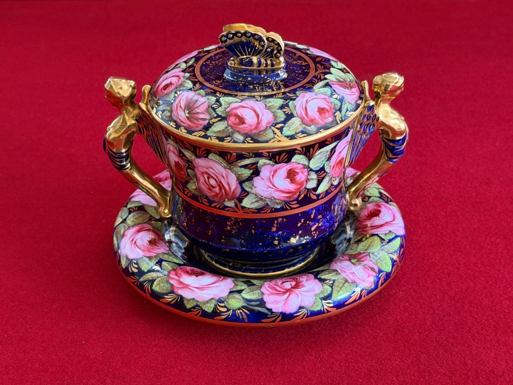 An antique Regency period porcelain covered chocolate cup with matching saucer, decorated in cobalt blue and hand-painted pink roses with winged figure handles and a butterfly finial.