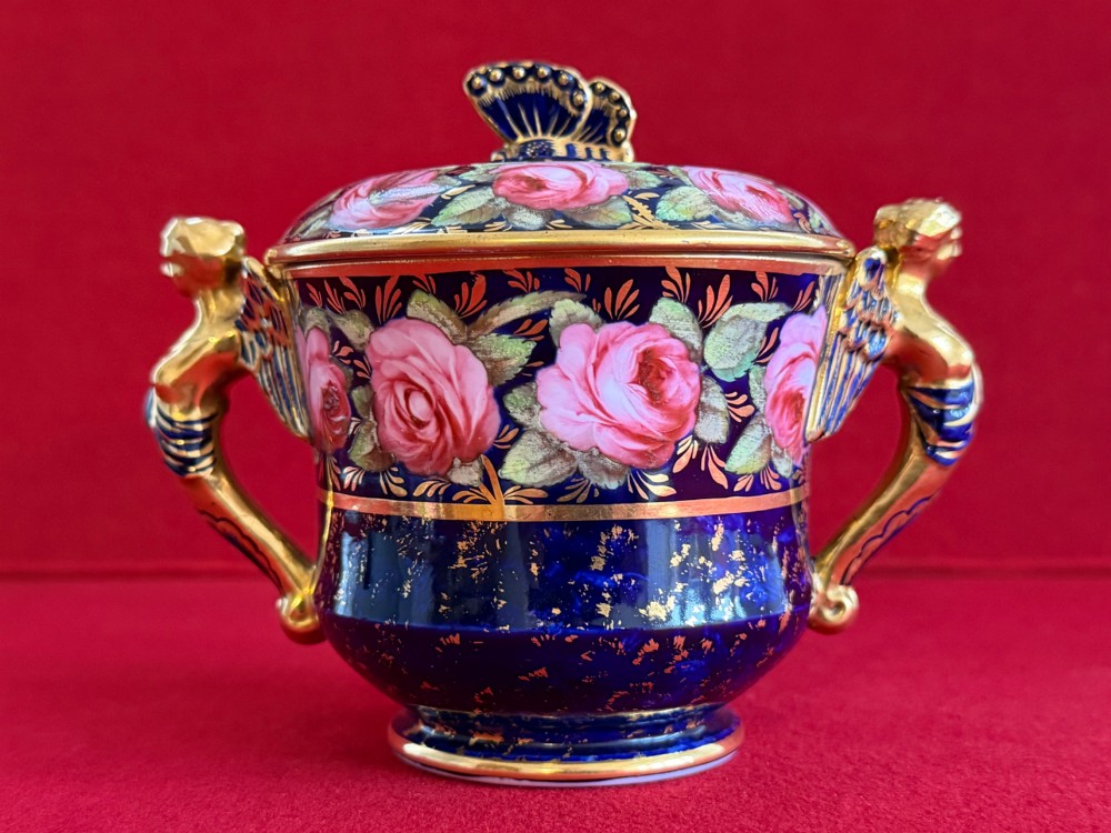 An antique Regency period porcelain covered chocolate cup with matching saucer, decorated in cobalt blue and hand-painted pink roses with winged figure handles and a butterfly finial. View II