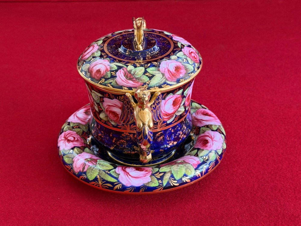An antique Regency period porcelain covered chocolate cup with matching saucer, decorated in cobalt blue and hand-painted pink roses with winged figure handles and a butterfly finial. View VII