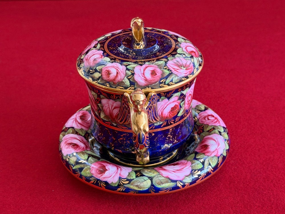 An antique Regency period porcelain covered chocolate cup with matching saucer, decorated in cobalt blue and hand-painted pink roses with winged figure handles and a butterfly finial. View IX