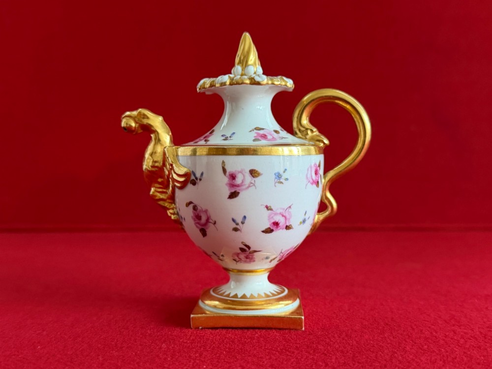 A Regency porcelain rose water sprinkler decorated with pink painted floral sprigs, a gilded spout, a scroll handle, and a square gilded plinth base.