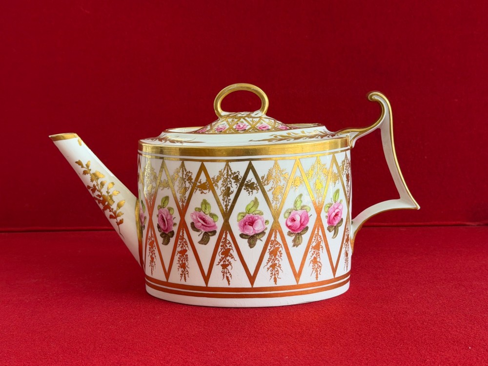 An antique oval white porcelain teapot decorated with hand-painted pink roses inside gilded diamond lozenges and gold rim details.