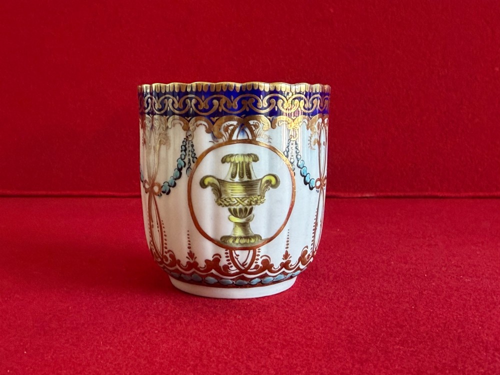 An 18th-century Worcester porcelain trio comprising a teacup, coffee cup, and saucer decorated with fluted moulding, blue swags, Classical urn motifs, and gold leaf gilding. View IX