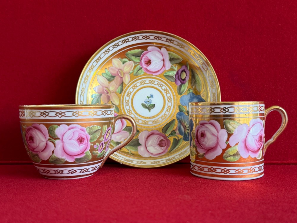A Regency John Rose Coalport porcelain trio comprising a teacup, coffee can, and matching saucer featuring hand-painted pink roses on a gold ground.