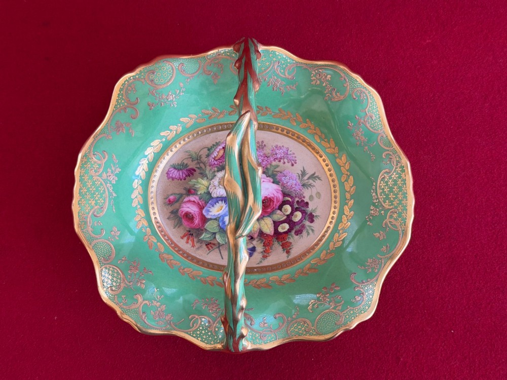 An antique green porcelain basket featuring a central hand-painted floral bouquet, gilt scrolling filigree, and a gilded branch handle.
