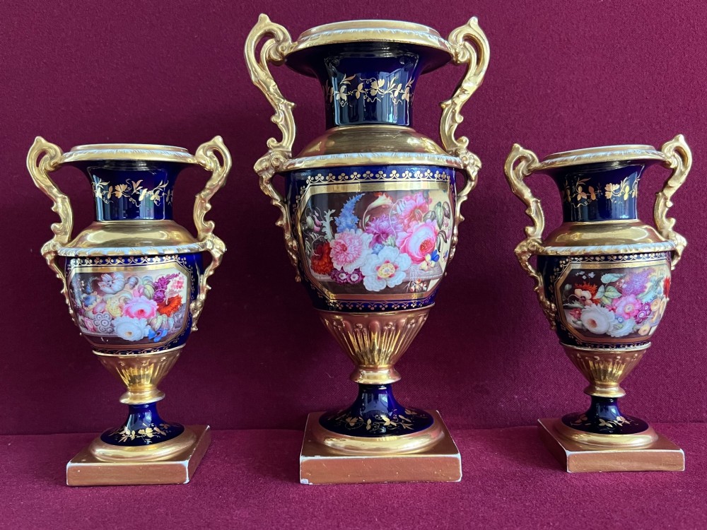 A three-piece set of cobalt blue and gilded Minton porcelain urn vases featuring hand-painted floral panels on square plinths.