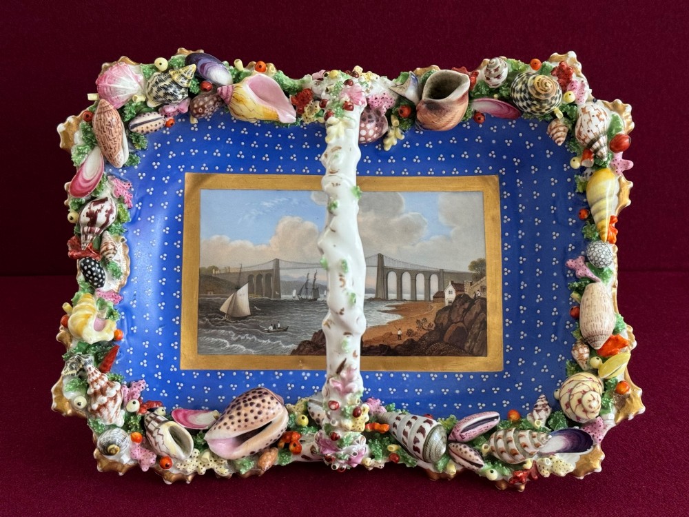 An antique Chamberlain Worcester porcelain basket featuring encrusted dimensional sea shells around the rim, an overhead central handle, and a hand-painted scene of the Menai Bridge set against a cobalt blue background.