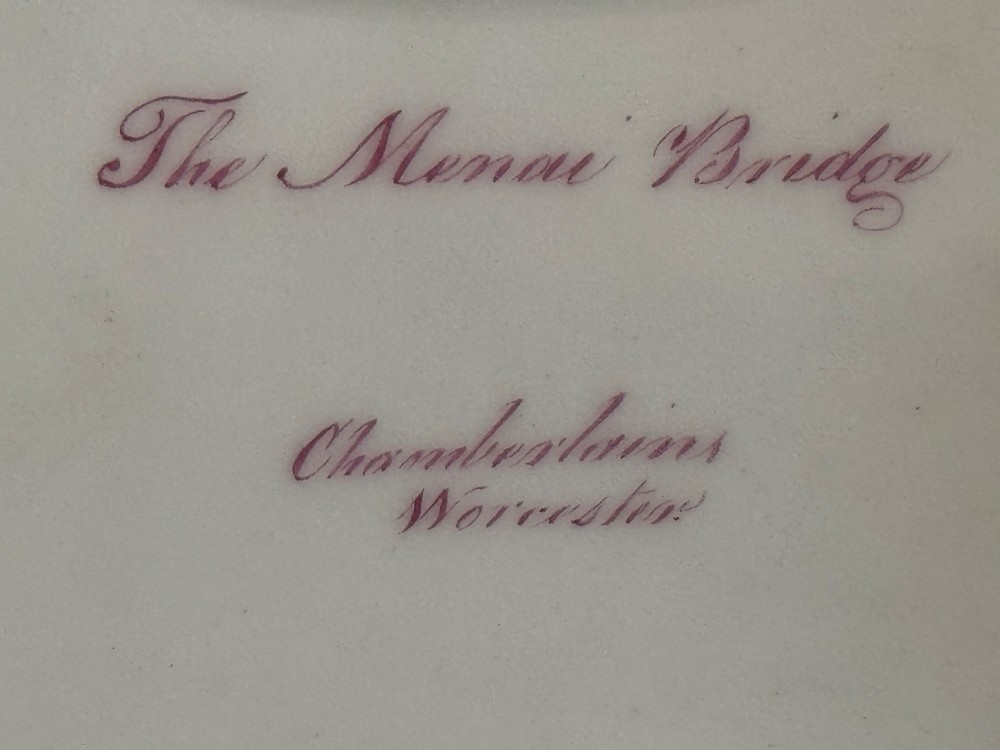An antique Chamberlain Worcester porcelain basket featuring encrusted dimensional sea shells around the rim, an overhead central handle, and a hand-painted scene of the Menai Bridge set against a cobalt blue background. View V
