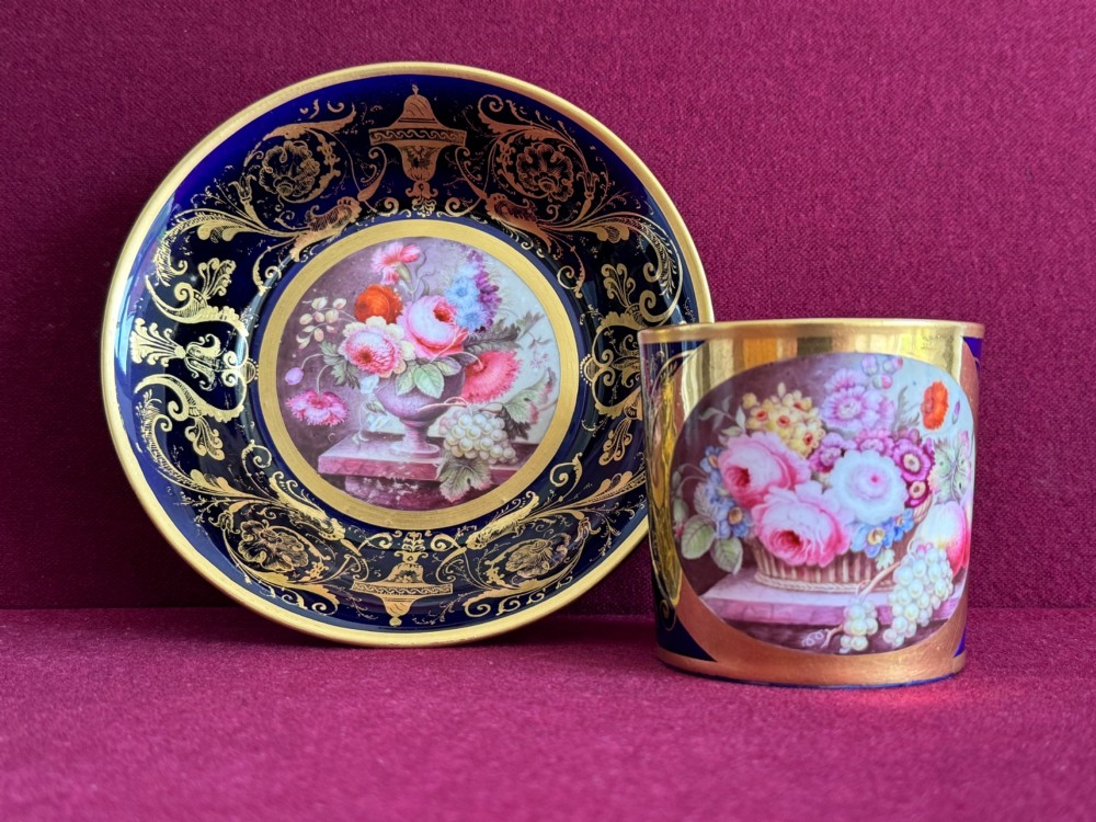 A cylindrical porcelain coffee can and saucer featuring deep cobalt blue grounds, elaborate gilt scrollwork, and central hand-painted floral still-life panels.