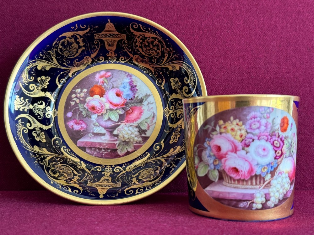 A cylindrical porcelain coffee can and saucer featuring deep cobalt blue grounds, elaborate gilt scrollwork, and central hand-painted floral still-life panels. View II