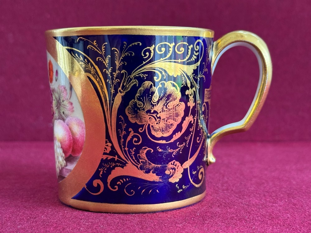 A cylindrical porcelain coffee can and saucer featuring deep cobalt blue grounds, elaborate gilt scrollwork, and central hand-painted floral still-life panels. View V