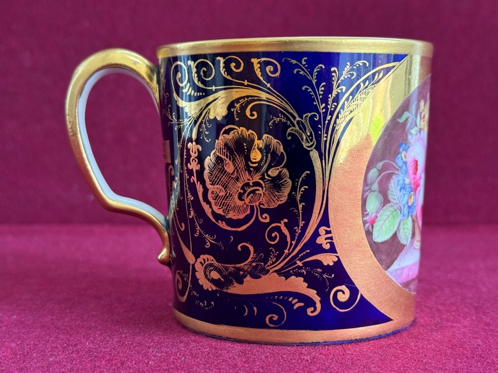 A cylindrical porcelain coffee can and saucer featuring deep cobalt blue grounds, elaborate gilt scrollwork, and central hand-painted floral still-life panels. View VI