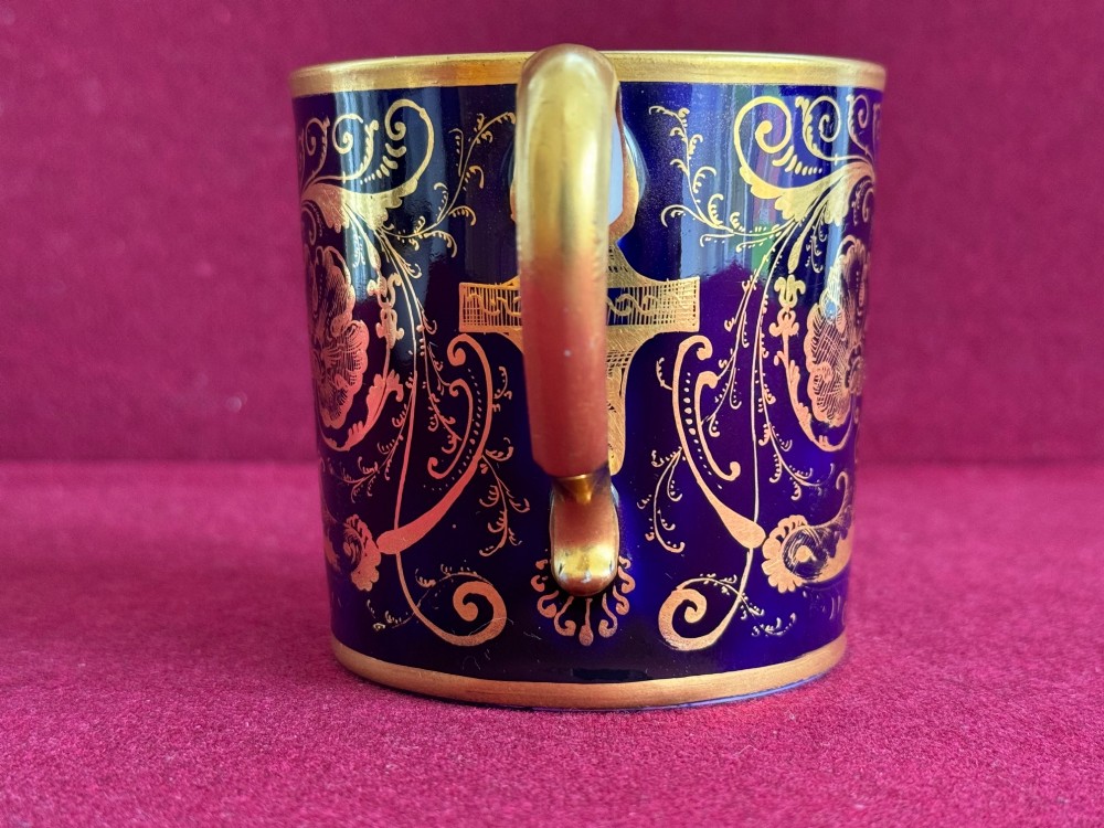 A cylindrical porcelain coffee can and saucer featuring deep cobalt blue grounds, elaborate gilt scrollwork, and central hand-painted floral still-life panels. View VII