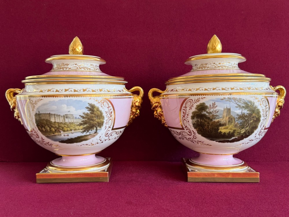 A pair of antique Regency porcelain ice pails featuring pink and white grounds, hand-painted landscape scenes, gilded lion mask handles, and pinecone finials.