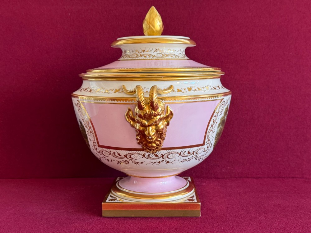 A pair of antique Regency porcelain ice pails featuring pink and white grounds, hand-painted landscape scenes, gilded lion mask handles, and pinecone finials. View V