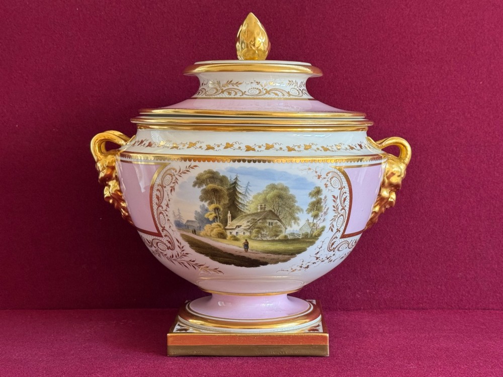 A pair of antique Regency porcelain ice pails featuring pink and white grounds, hand-painted landscape scenes, gilded lion mask handles, and pinecone finials. View X
