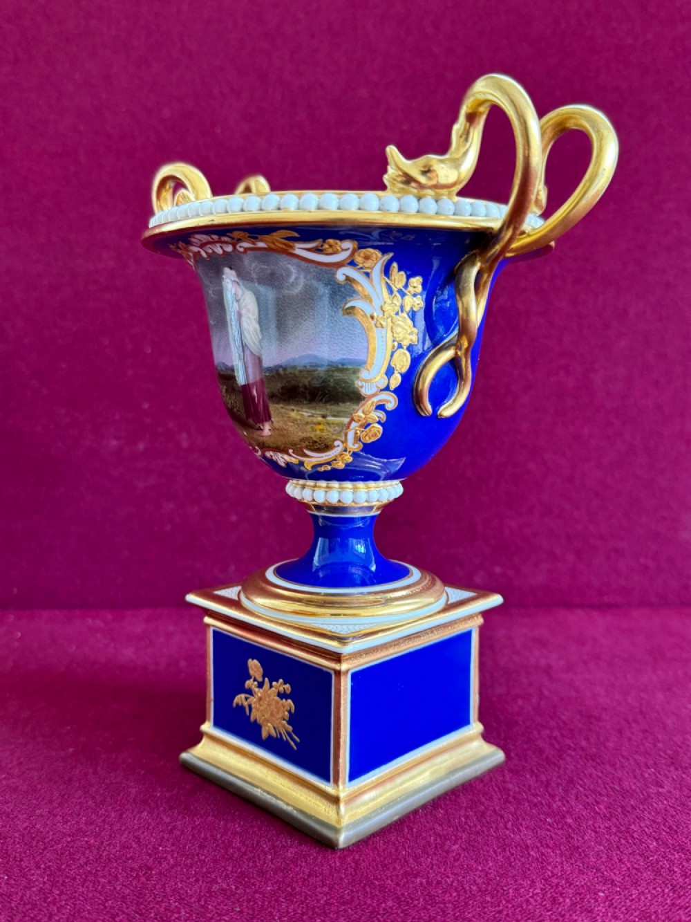 A Regency cobalt blue and gilded porcelain urn featuring a central hand-painted panel of a standing figure in a landscape, twin scroll handles, beaded borders, and a square pedestal base. View II