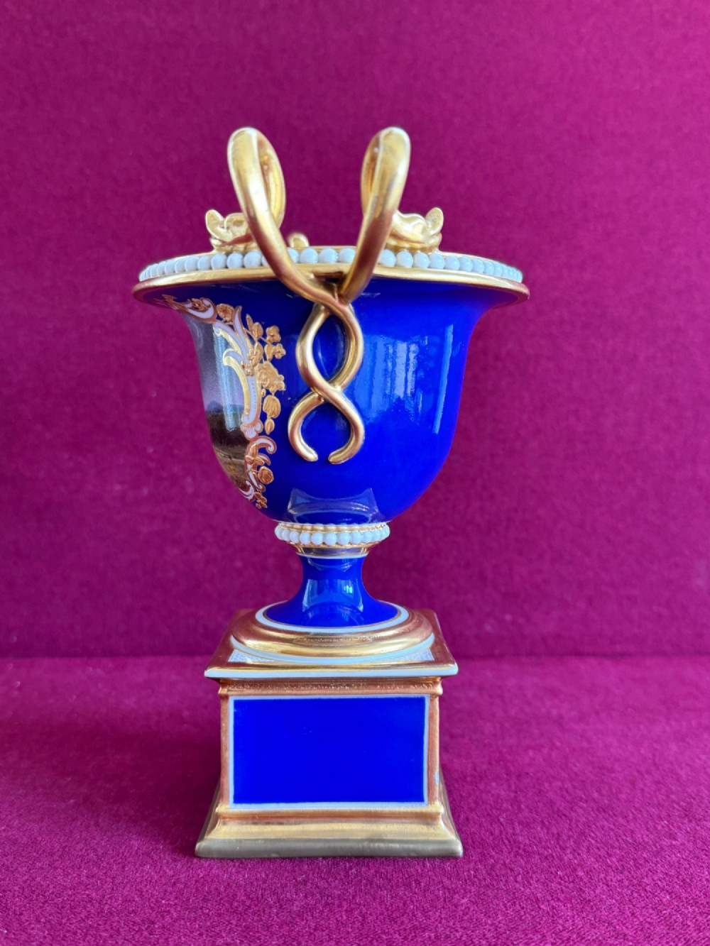 A Regency cobalt blue and gilded porcelain urn featuring a central hand-painted panel of a standing figure in a landscape, twin scroll handles, beaded borders, and a square pedestal base. View III