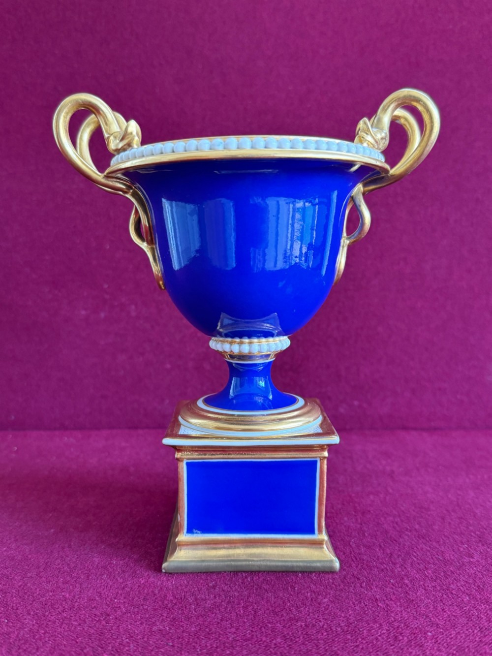 A Regency cobalt blue and gilded porcelain urn featuring a central hand-painted panel of a standing figure in a landscape, twin scroll handles, beaded borders, and a square pedestal base. View IV