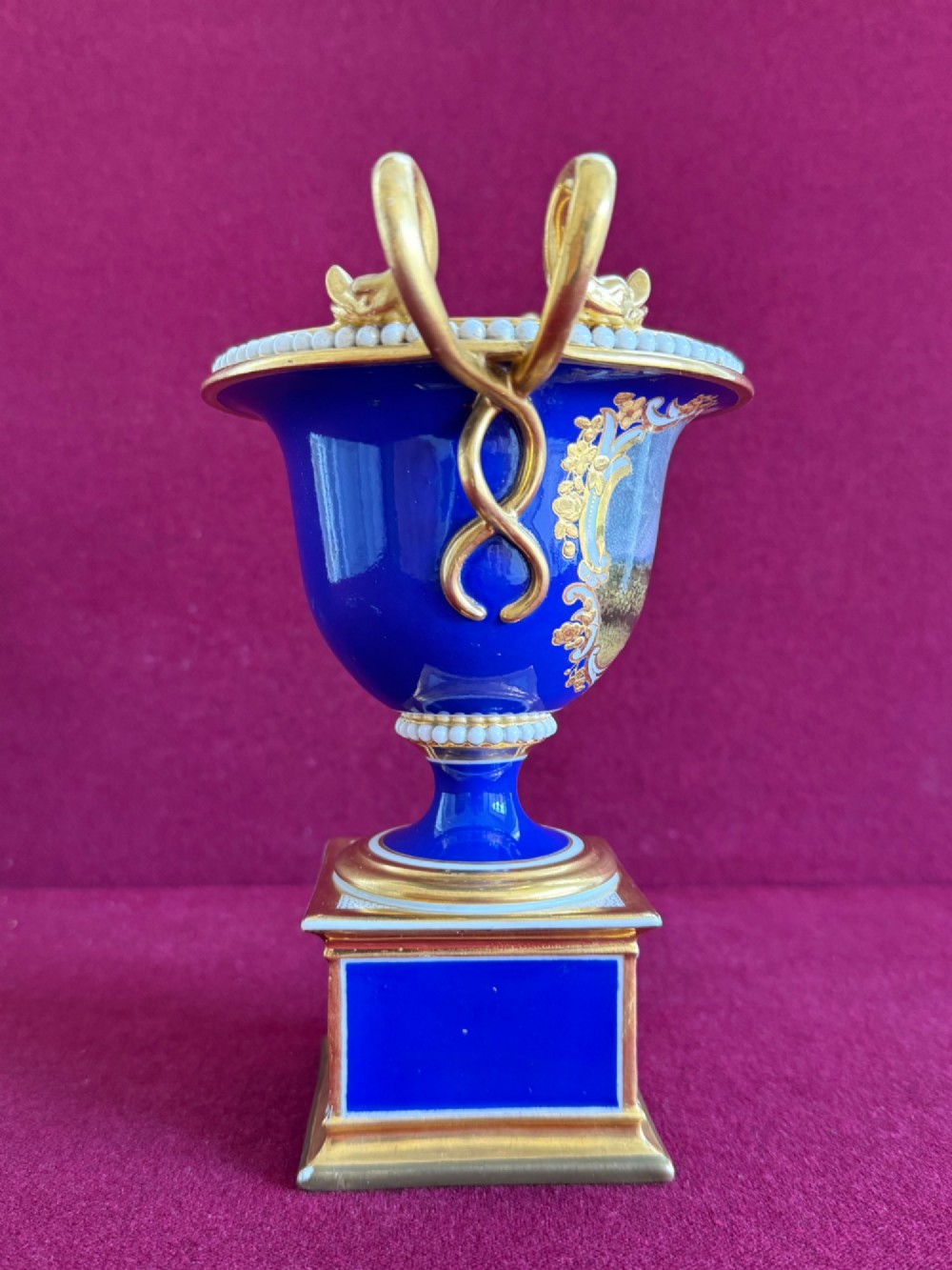 A Regency cobalt blue and gilded porcelain urn featuring a central hand-painted panel of a standing figure in a landscape, twin scroll handles, beaded borders, and a square pedestal base. View V
