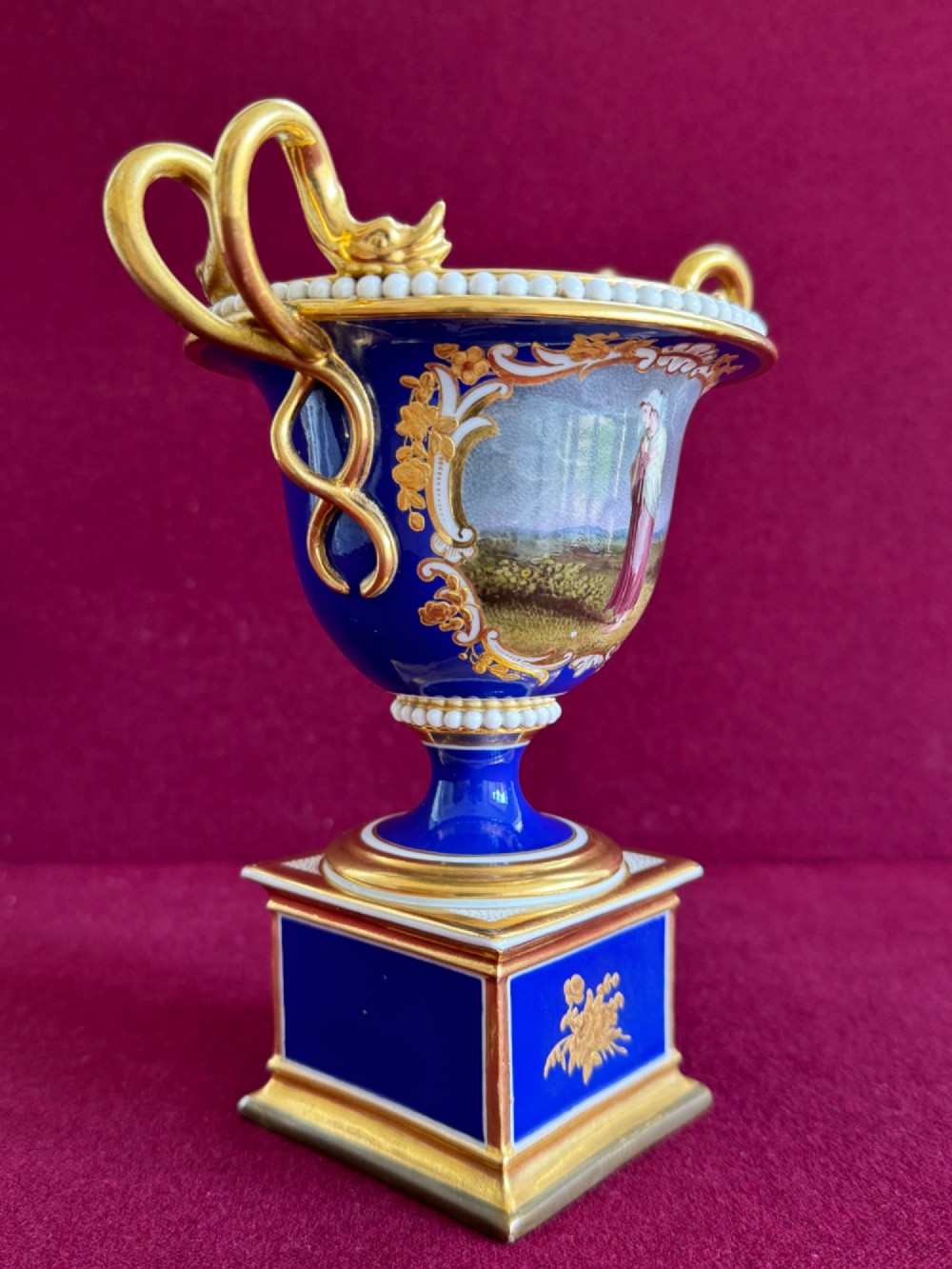 A Regency cobalt blue and gilded porcelain urn featuring a central hand-painted panel of a standing figure in a landscape, twin scroll handles, beaded borders, and a square pedestal base. View VI