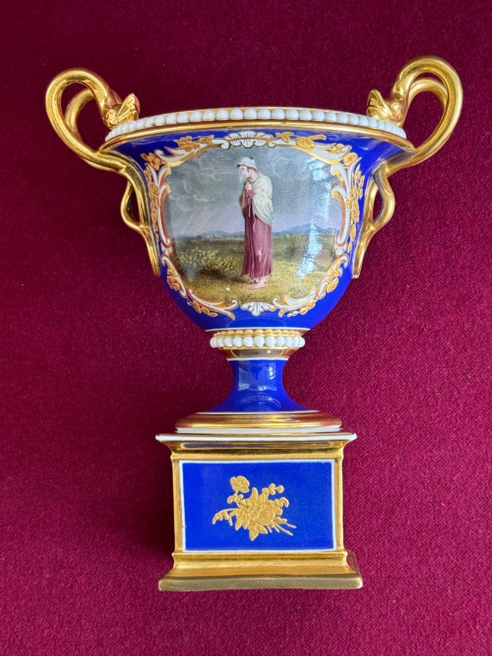 A Regency cobalt blue and gilded porcelain urn featuring a central hand-painted panel of a standing figure in a landscape, twin scroll handles, beaded borders, and a square pedestal base. View VII