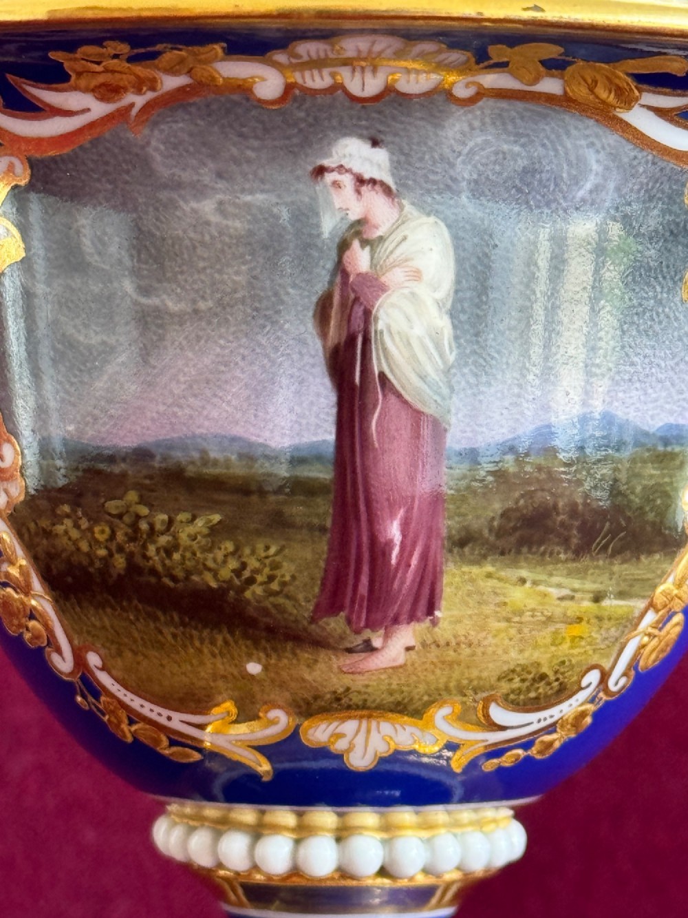 A Regency cobalt blue and gilded porcelain urn featuring a central hand-painted panel of a standing figure in a landscape, twin scroll handles, beaded borders, and a square pedestal base. View XI