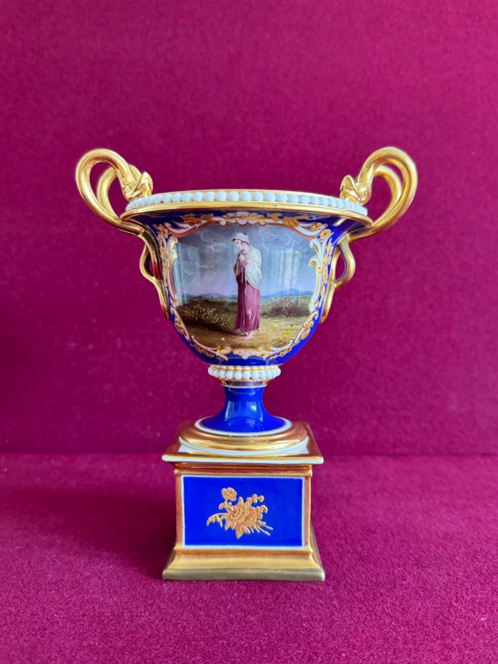 A Regency cobalt blue and gilded porcelain urn featuring a central hand-painted panel of a standing figure in a landscape, twin scroll handles, beaded borders, and a square pedestal base. View XII