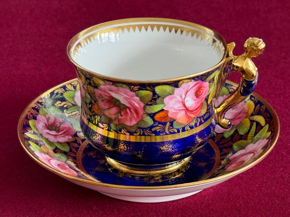 An antique Regency period Spode porcelain teacup and saucer decorated with hand-painted pink roses, cobalt blue ground, gold leaf gilding, and a gilded figural handle.