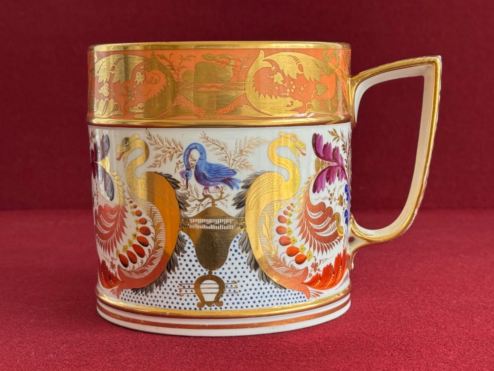 A cylindrical Regency period Derby porcelain porter mug decorated with hand-painted bird motifs, gold leaf foliage, an orange top border, and an angular gilded handle.