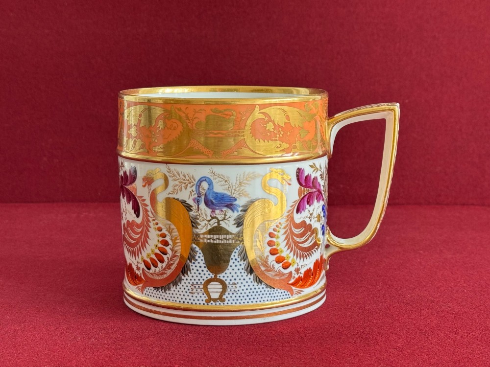 A cylindrical Regency period Derby porcelain porter mug decorated with hand-painted bird motifs, gold leaf foliage, an orange top border, and an angular gilded handle. View II